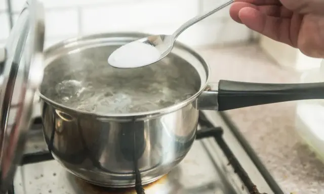 En sked med salt tillsätts i kokande vatten. En del av locket är upplyft. En burk med salt syns i bakgrunden, redo för egen resorb.