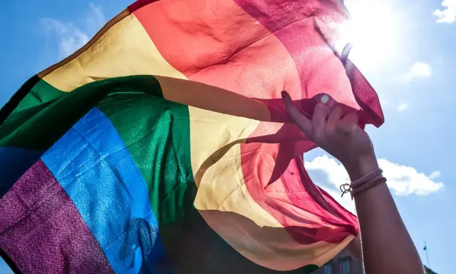 En hand håller en regnbågsflagga mot en solig himmel. En symbol för att **run for pride** och kärlek.