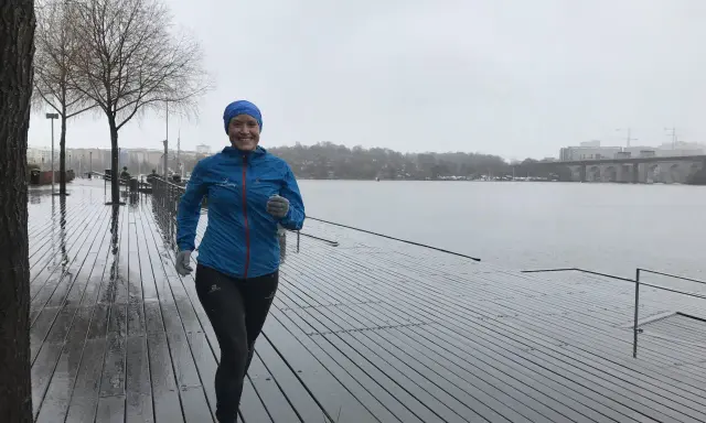 Glad kvinna med blå jacka och mössa springer i regn längs en träbrygga vid en sjö.