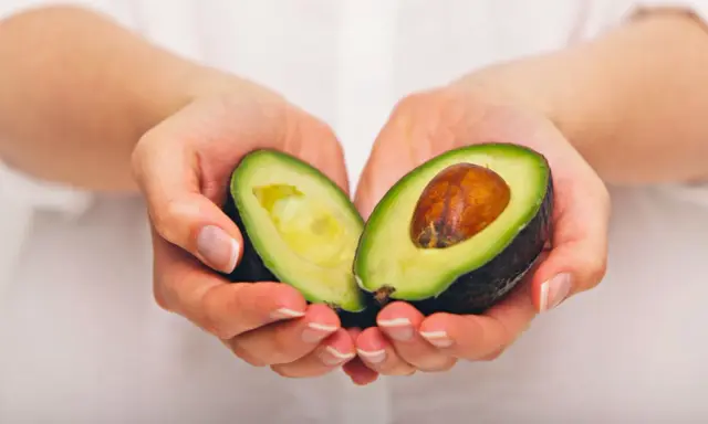 Händerna håller en avokado, delad på mitten, som visar dess gröna kött och bruna kärna. En näringsrik källa till avokado vitaminer.