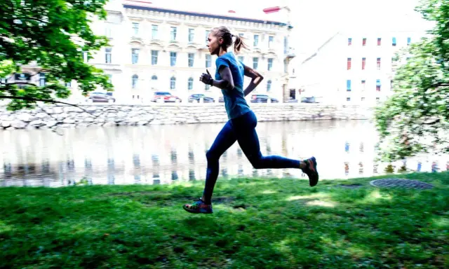 En kvinna joggar långsamt eller går snabbt längs en gräsmatta vid en flod, med byggnader i bakgrunden.