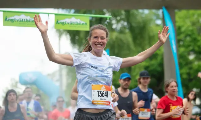 Glad kvinna med armarna uppe vid målgången i ett maraton. En lycklig löpare som njuter av sin maraton tid.