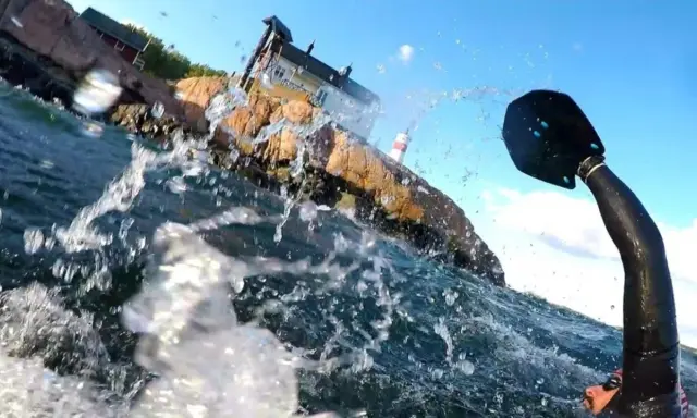 Vattenstänk och en paddel i förgrunden. I bakgrunden en klippa med hus och ett litet fyrhus. En del av en farleden swimrun-deltagare syns.