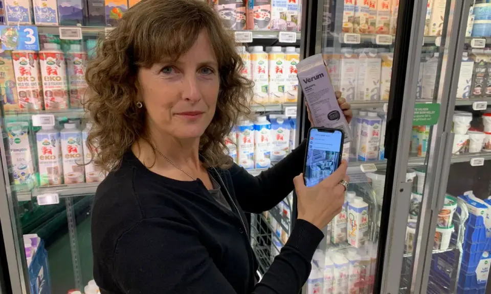 Kvinna håller upp en mjölkförpackning och en mobiltelefon, visar hur man kollar vad som innehåller socker. Kylskåp med mejeriprodukter i bakgrunden.
