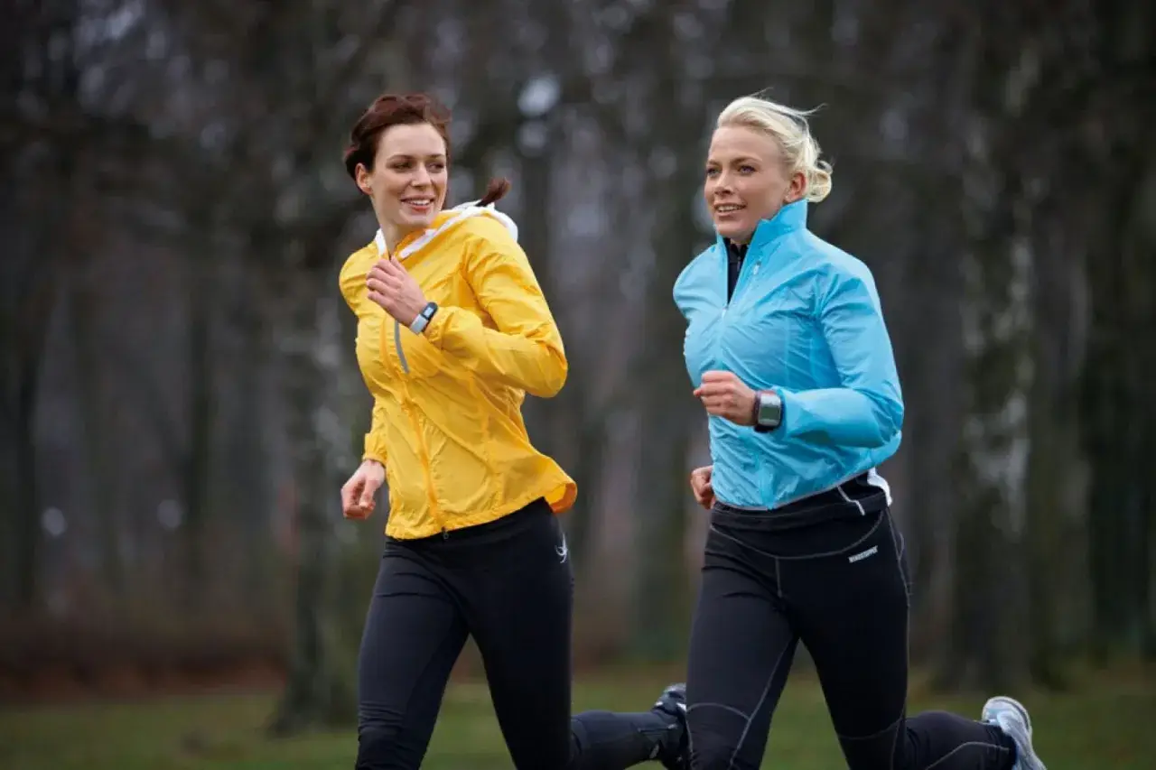 Två kvinnor springer i parken, förberedda för ett halv maraton. Den ena bär en gul jacka, den andra en blå.