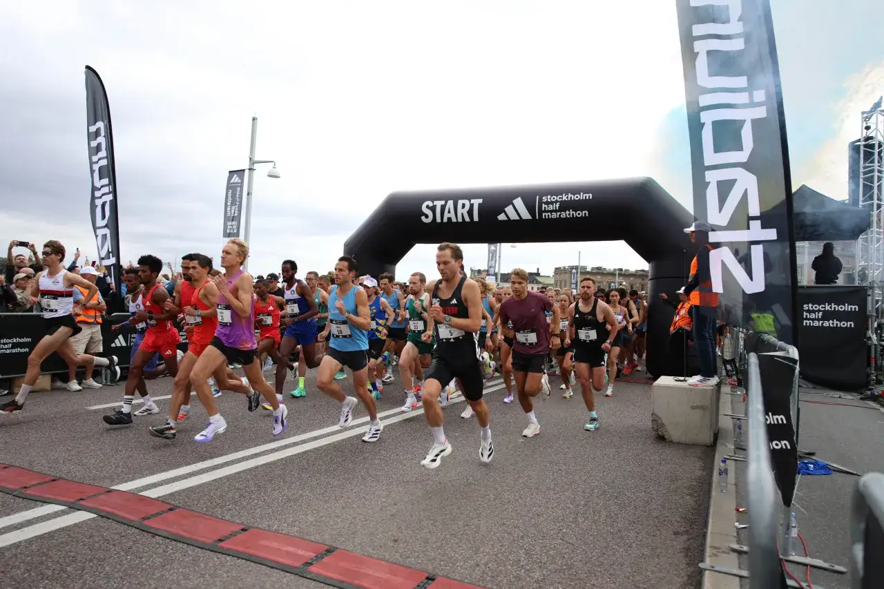 Löpare springer mot startlinjen för Stockholm Halvmaraton. En stor svart båge markerar starten, och deltagarna är redo att kämpa för bästa halvmaraton tid.