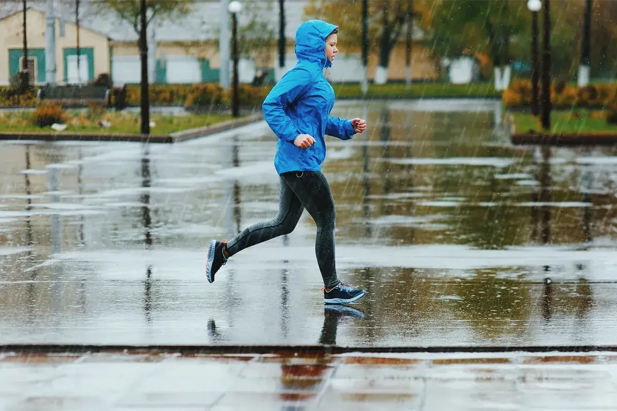 En person i blå jacka med huva springer i regnet på en blöt trottoar.