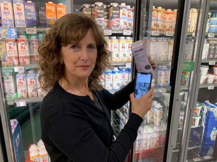 Kvinna håller upp en mjölkförpackning och en mobiltelefon, visar hur man kollar vad som innehåller socker. Kylskåp med mejeriprodukter i bakgrunden.
